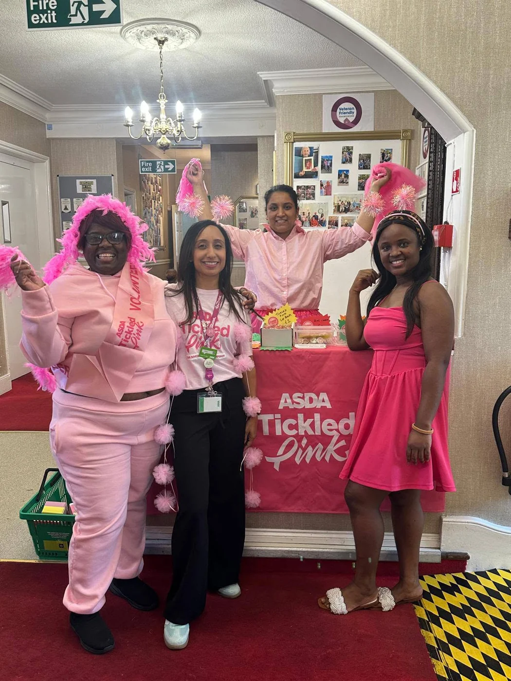 Four people dressed in pink, wearing Asda Tickled Pink sashes and feather boas, standing beside the pink stall in the entrance hall of the care home.