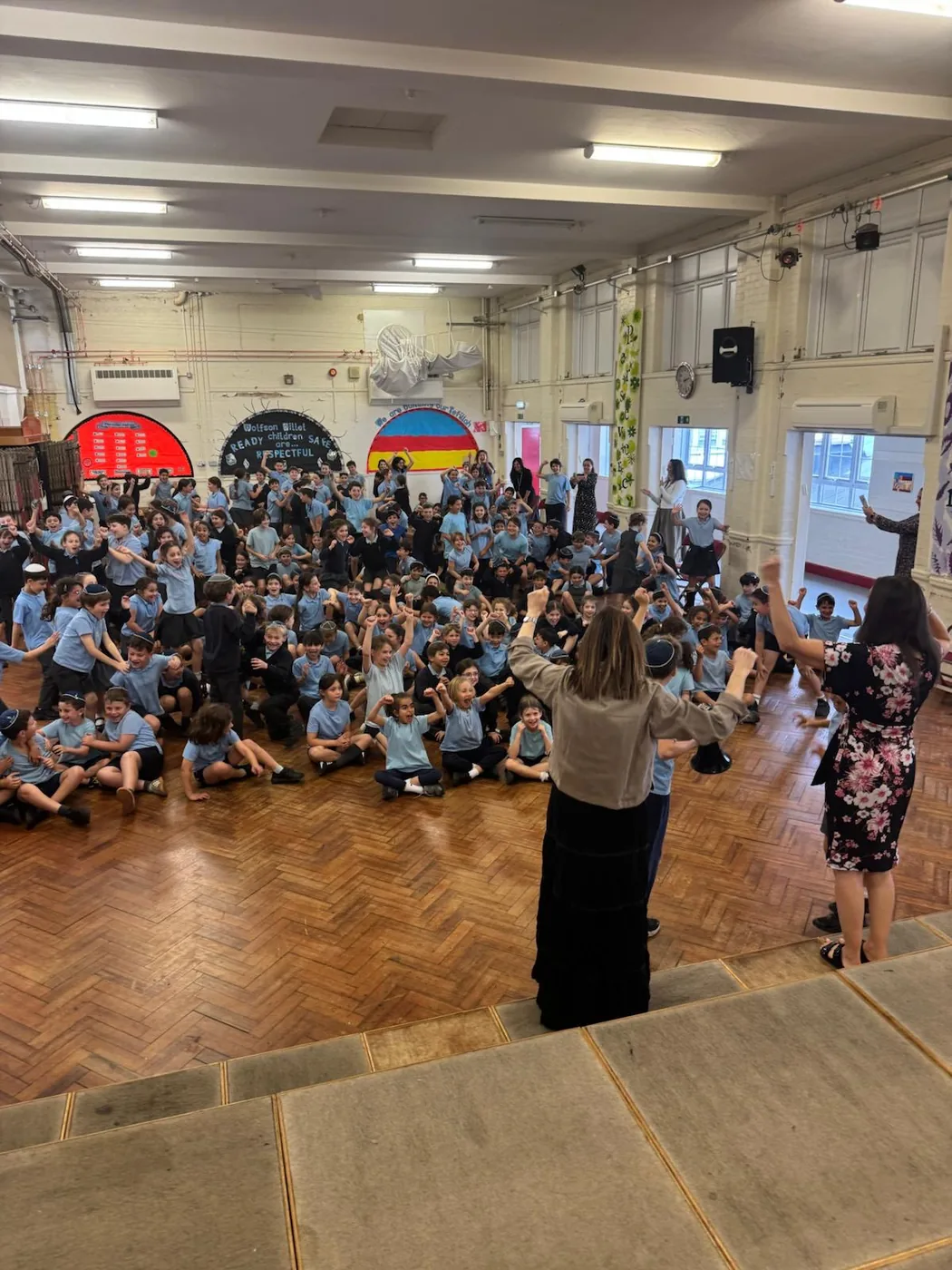 Pupils and staff filling the school hall, arms raised, in front of a wall display reading Wolfson Hillel children safe ready respectful